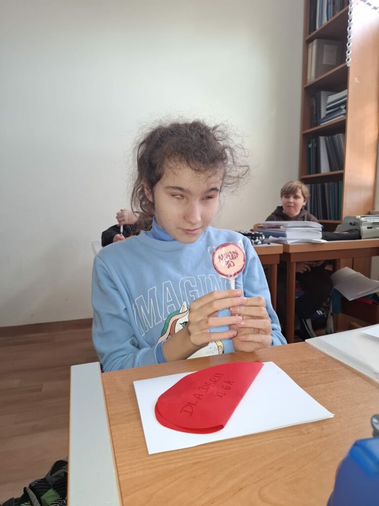 Uczennica w błękitnej bluzie siedzi przy ławce, trzymając walentynkowego lizaka z napisem „KOCHAM CIĘ”, obok leżącej na papierze czerwonej koperty z dedykacją.