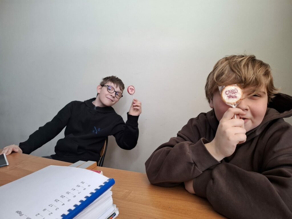 Dwóch uczniów siedzi przy szkolnej ławce, prezentując do aparatu walentynkowe lizaki, z których jeden ma napis „CAŁUJ MNIE”.