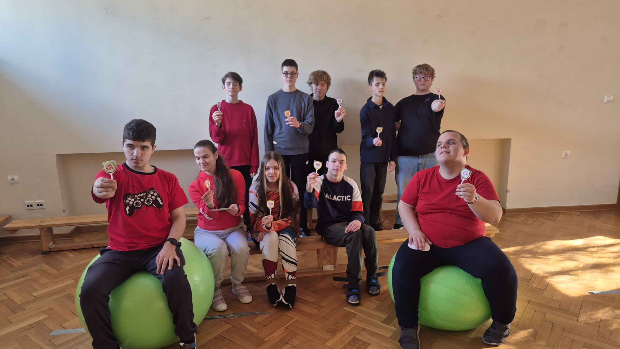 Grupa starszych uczniów pozuje w sali gimnastycznej, siedząc na zielonych piłkach rehabilitacyjnych i ławkach, trzymając w dłoniach walentynkowe lizaki otrzymane od Pani Dyrektor.
