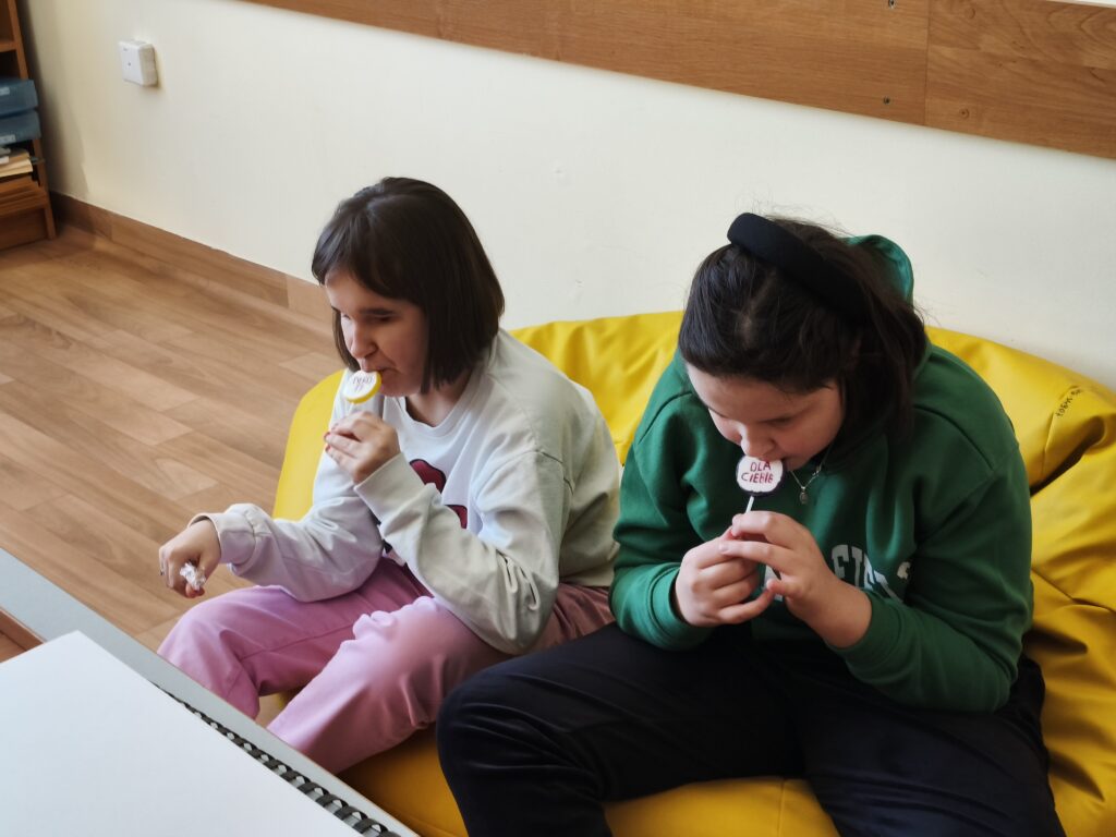 Dwie dziewczynki siedzą na żółtej pufie i z radością jedzą walentynkowe lizaki z napisem „Dla Ciebie”, które otrzymały w ramach szkolnego świętowania.