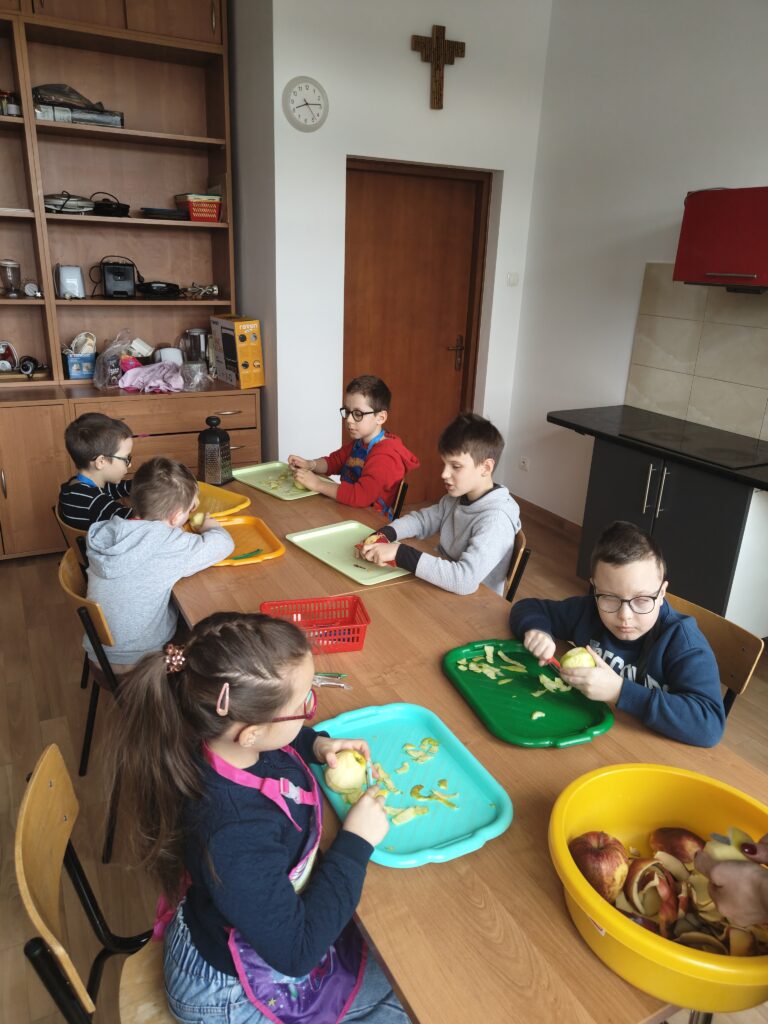 Na zdjęciu grupa skupionych uczniów siedzi przy długim stole, wspólnie obierając jabłka na kolorowych taca (zielonych i pomarańczowych). Dzieci z zaangażowaniem używają obierek, przygotowując owoce, które staną się głównym składnikiem brownie