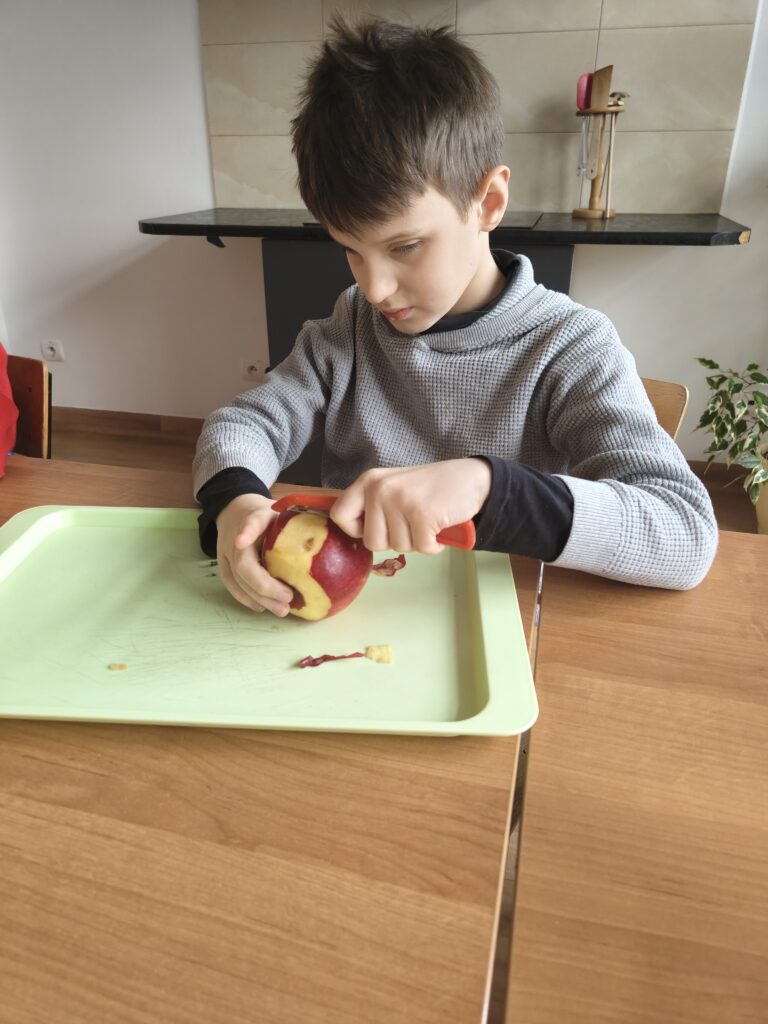 Chłopiec w szarym swetrze uważnie posługuje się czerwoną obieraczką, zdejmując skórkę z dużego czerwonego jabłka nad jasnozieloną tacką.