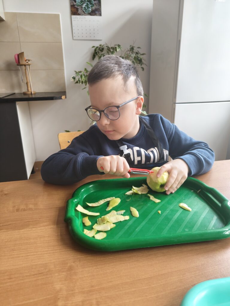 Na zdjęciu widzimy chłopca w okularach i granatowej bluzie, który z dużym skupieniem obiera zielone jabłko nad ciemnozieloną tacką, używając do tego czerwonej obieraczki.