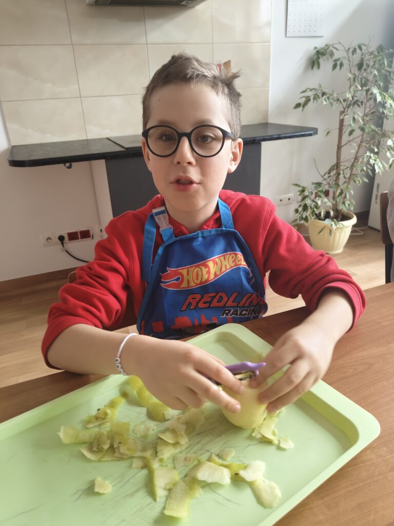 Na zdjęciu widzimy chłopca w okularach i niebieskim fartuszku z motywem "Hot Wheels", który siedzi przy jasnym stole i z zaangażowaniem obiera jabłko nad seledynową tacką.
