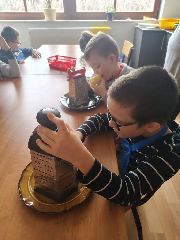 Na zbliżeniu widać dwóch uczniów zaangażowanych w tarcie owoców, gdzie jeden z nich oburącz trzyma dużą tarkę na talerzu, a drugi w tym samym czasie ze smakiem zajada kawałek obranego jabłka.