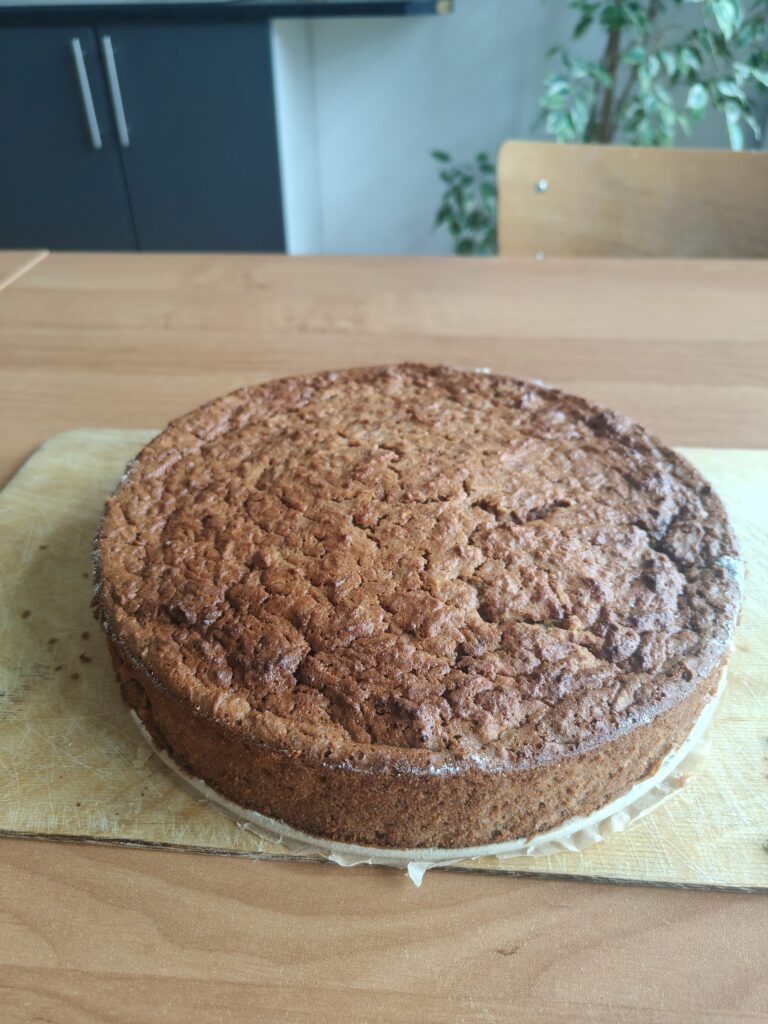 To zdjęcie przedstawia upieczone, okrągłe brownie jabłkowe o rumianej, lekko spękanej skórce, które spoczywa na drewnianej desce do krojenia, będąc dumnym efektem wspólnej pracy uczniów klas 1b i 3 podczas zajęć kulinarnych.