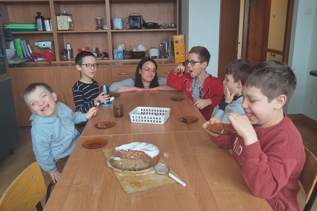 Na zdjęciu grupa radosnych uczniów i opiekunka siedzą wspólnie przy długim drewnianym stole, uśmiechając się i z apetytem degustując własnoręcznie upieczone, posypane cukrem pudrem brownie jabłkowe, co wieńczy ich pełne współpracy zajęcia kulinarne.