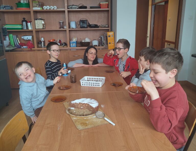 Na zdjęciu grupa radosnych uczniów i opiekunka siedzą wspólnie przy długim drewnianym stole, uśmiechając się i z apetytem degustując własnoręcznie upieczone, posypane cukrem pudrem brownie jabłkowe, co wieńczy ich pełne współpracy zajęcia kulinarne.