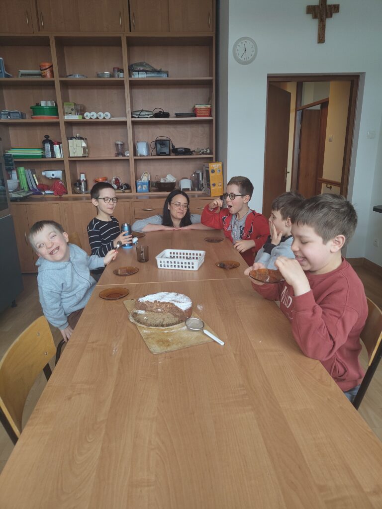 Na zdjęciu grupa radosnych uczniów i opiekunka siedzą wspólnie przy długim drewnianym stole, uśmiechając się i z apetytem degustując własnoręcznie upieczone, posypane cukrem pudrem brownie jabłkowe, co wieńczy ich pełne współpracy zajęcia kulinarne.