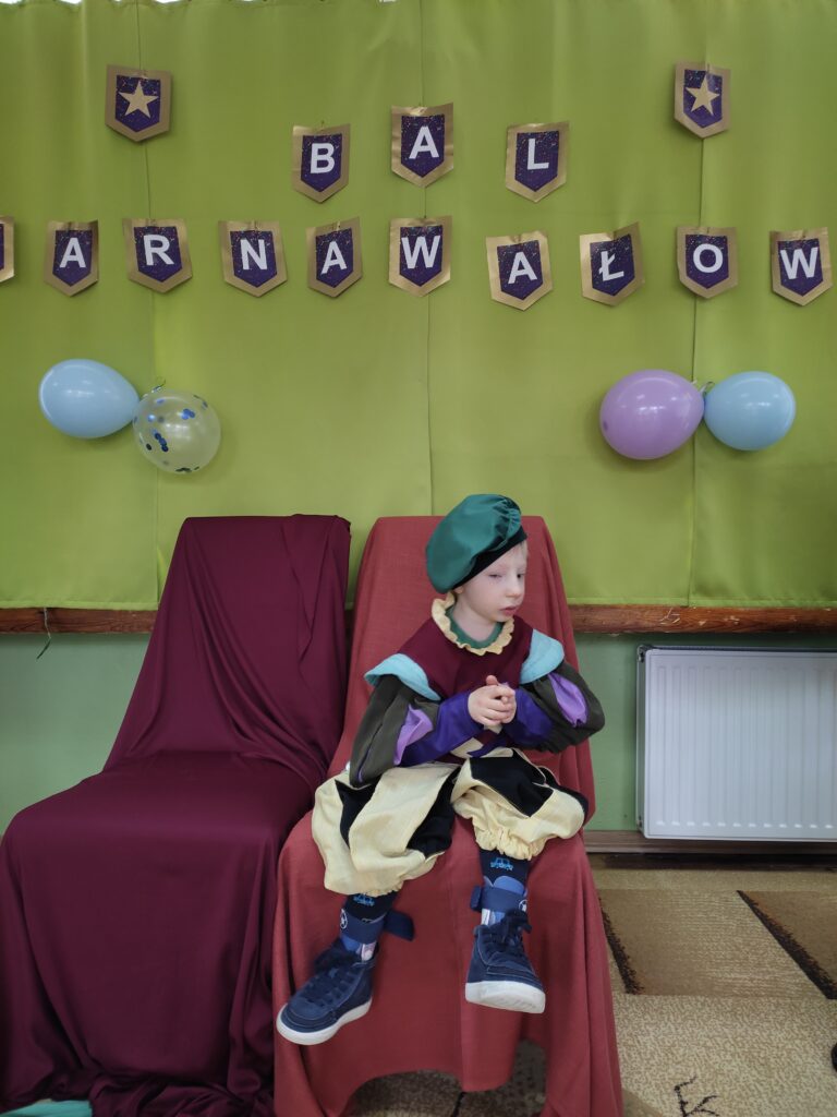 Chłopiec przebrany za dworzanina siedzi na krześle w tle napis Bal Karnawałowy.