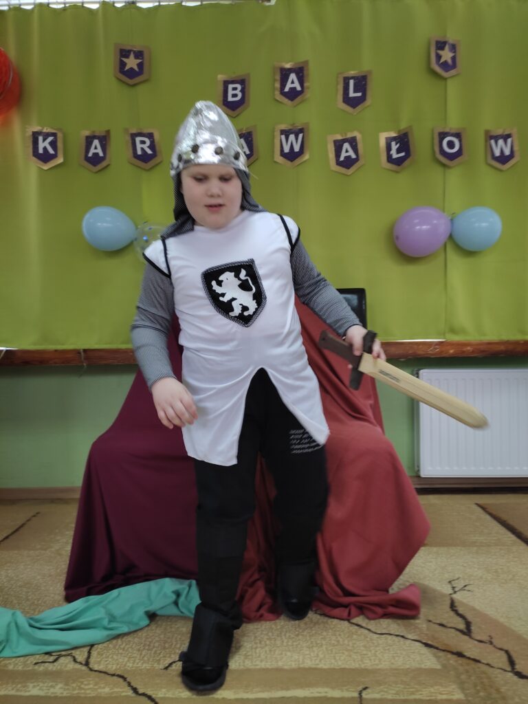 Chłopiec przebrany za rycerza stoi z mieczem w dłoni w tle napis Bal Karnawałowy.