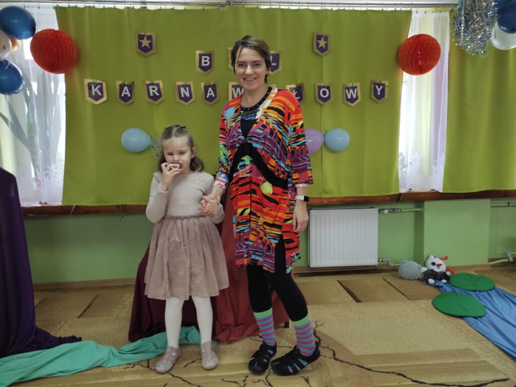 Nauczycielka przebrana za Stańczyka z dziewczynką przebraną za królewnę w tle napis Bal Karnawałowy.