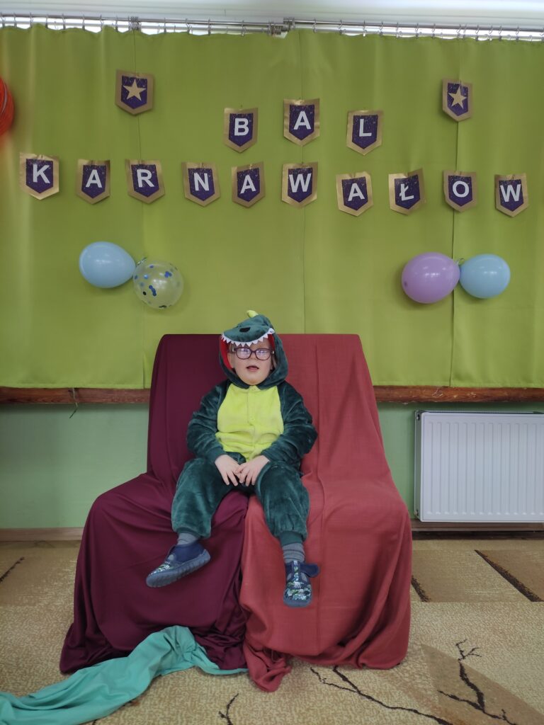 Chłopiec przebrany za smoka siedzi na krześle w tle napis Bal Karnawałowy.