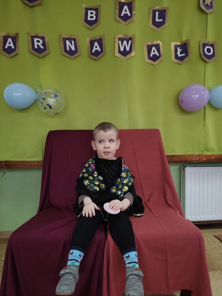 Chłopiec przebrany za herolda siedzi na krześle tle napis Bal Karnawałowy.