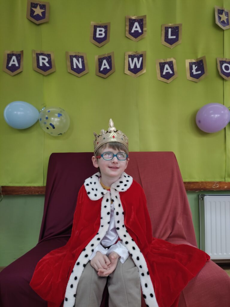 Chłopiec przebrany za króla siedzi na krześle w tle napis Bal Karnawałowy.