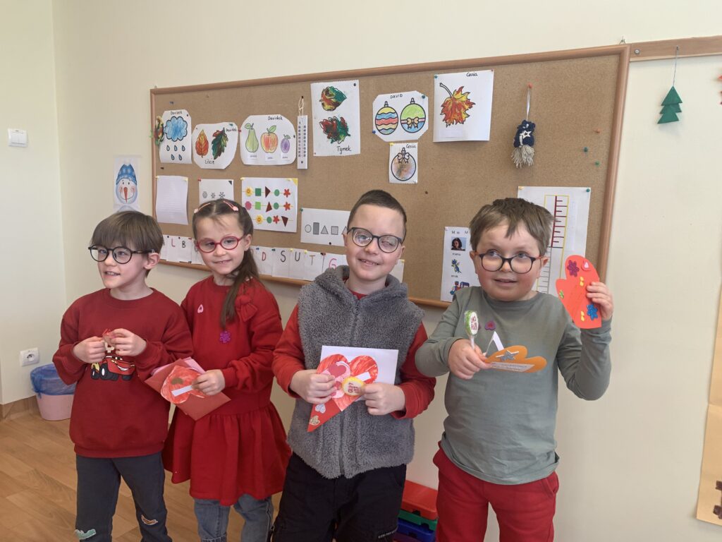 Czworo uśmiechniętych młodych uczniów stoi na tle tablicy z pracami plastycznymi, dumnie prezentując własnoręcznie wykonane, kolorowe serca oraz walentynkowe upominki.