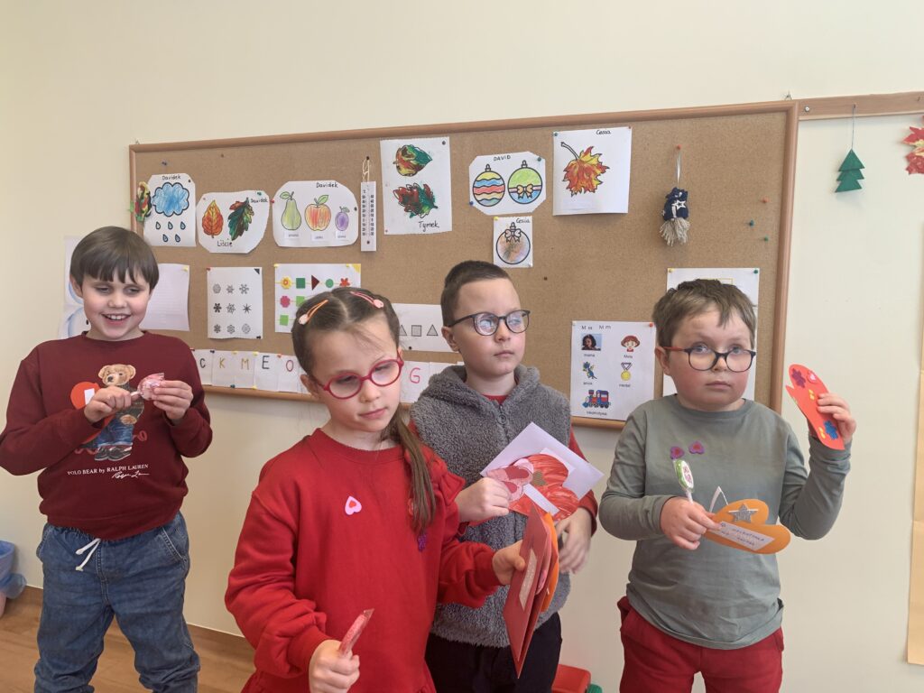 Czworo uśmiechniętych dzieci w okularach stoi na tle tablicy z pracami plastycznymi, dumnie prezentując własnoręcznie wykonane, kolorowe serca oraz walentynkowe upominki.