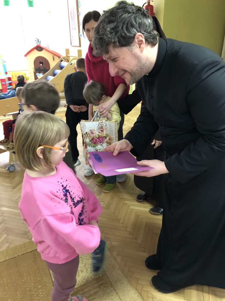 Ksiądz z uśmiechem trzyma w ręku laurkę, przed nim dziewczynka.