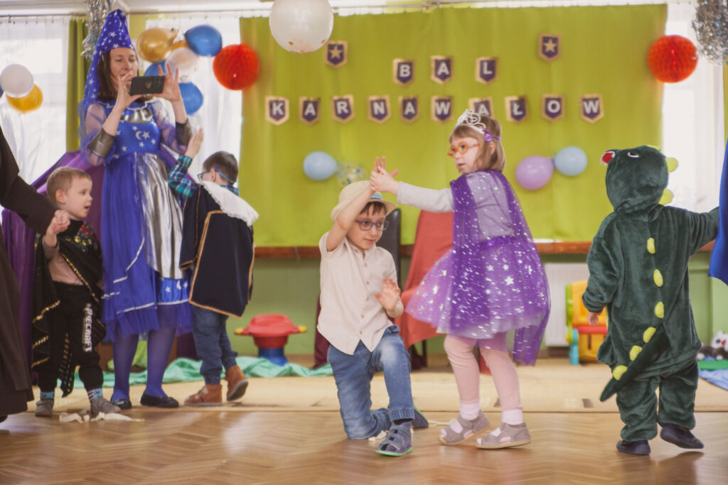 Chłopiec klęczy na jednym kolanie ma podniesioną rękę dziewczynka trzyma go za dłoń i tańczy dookoła . W tle inne dzieci.