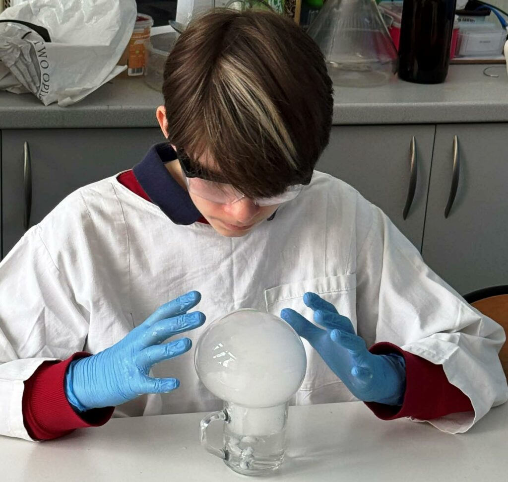 Osoba w białym fartuchu laboratoryjnym i niebieskich rękawiczkach trzyma dłonie wokół dużej, białej bańki wypełnionej mgłą, która uformowała się na szklanym naczyniu z wodą i suchym lodem.