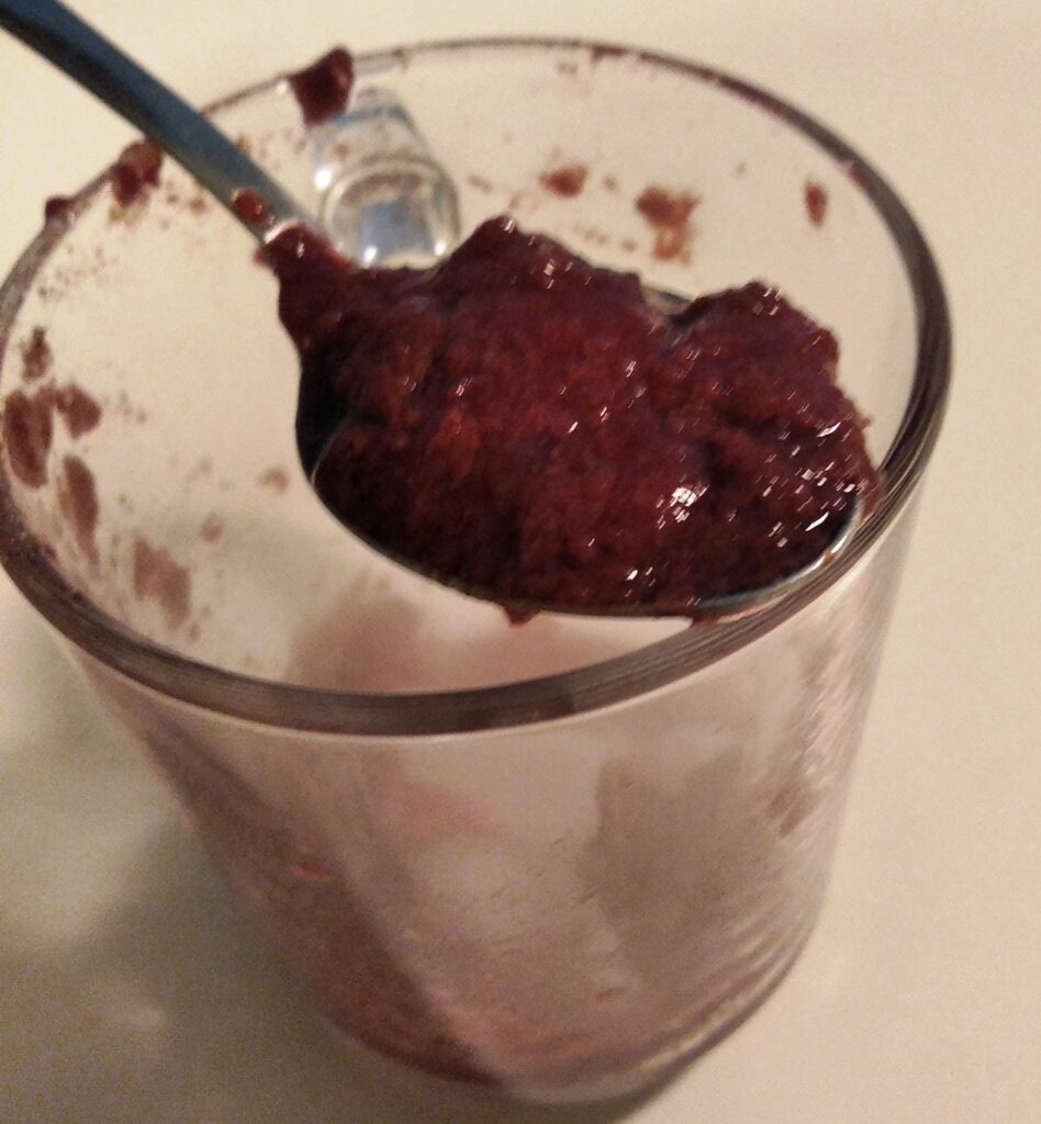 Zbliżenie na metalową łyżkę, na której znajduje się porcja gotowego deseru o gęstej konsystencji i głębokim, bordowym kolorze. Łyżka spoczywa na krawędzi szklanego naczynia, w którym widać resztę przygotowanego przysmaku.