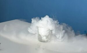 Z przezroczystej szklanki stojącej na białym stole wydobywa się gęsta, kłębiąca się biała mgła, która przelewa się przez krawędzie naczynia i nisko ściele się po powierzchni blatu. W tle widoczna jest jednolita niebieska ściana.