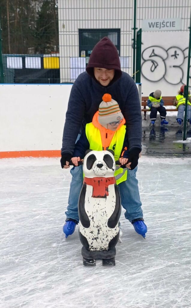 Chłopiec stoi na lodowisku, przed nim pchacz -panda, za nim opiekun