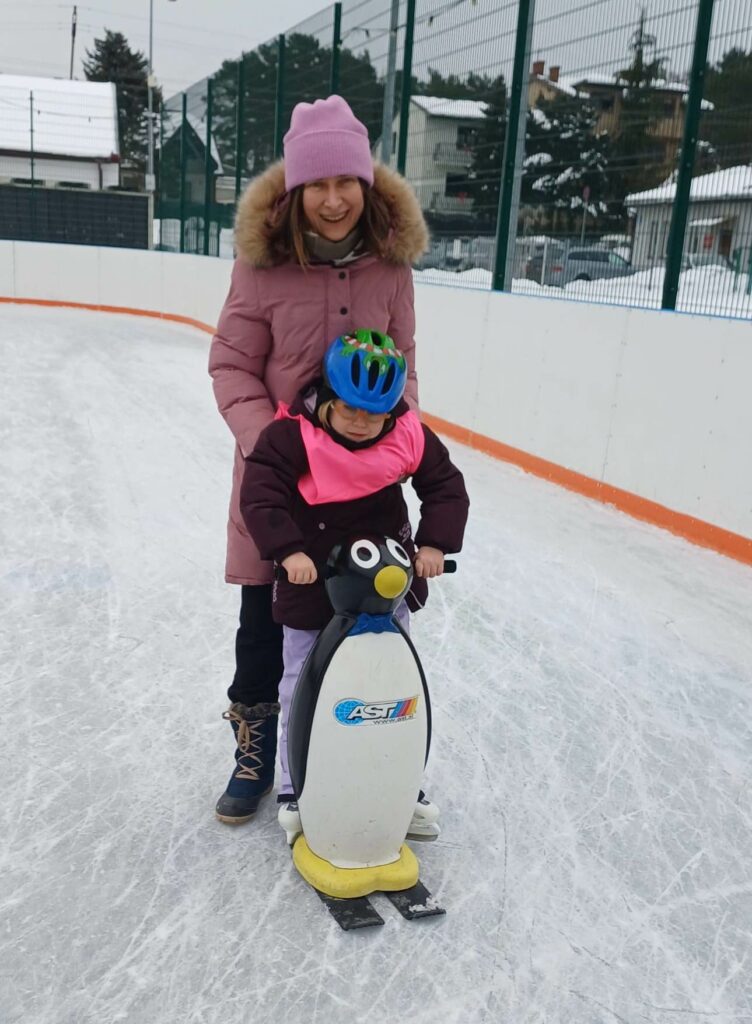 Dziewczynka w kasku i łyżwach stoi na lodowisku, przed nią pchacz - pingwin, za nią opiekunka,