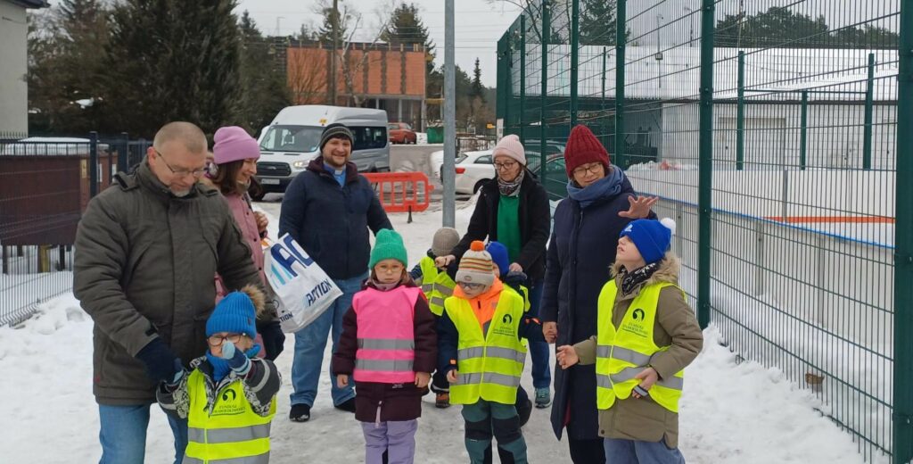 Dzieci w kamizelkach odblaskowych i opiekunowie stoją przy ogrodzeniu lodowiska