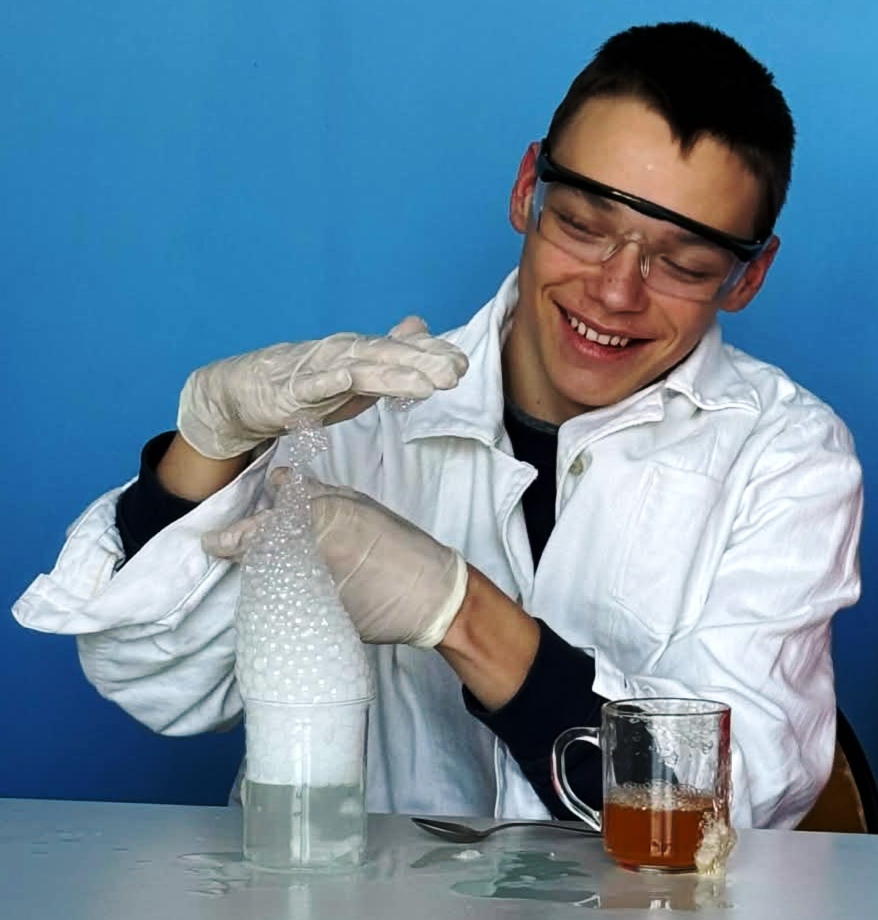 Uśmiechnięty uczeń w okularach ochronnych i białym fartuchu obserwuje wysoką kolumnę białej, gęstej piany, która wypływa ze szklanki z wodą. Eksperymentator ma na dłoniach jasne rękawiczki i wykonuje gest, jakby formował pianę, a obok na blacie stoi drugie naczynie z brązowawym płynem.