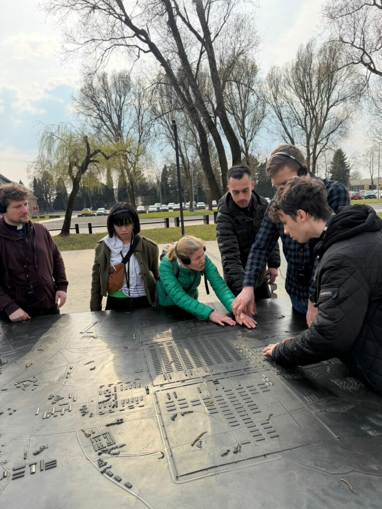 Podczas zajęć edukacyjnych w Muzeum Auschwitz-Birkenau maturzyści wraz z opiekunami w skupieniu analizują makietę terenu obozu, co było elementem poruszającej lekcji historii poświęconej pamięci o ofiarach i refleksji nad godnością ludzkiego życia.