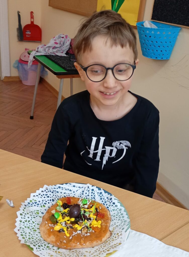 Uśmiechnięty chłopiec siedzi a przed nim na stoliku leży ozdobiony mazurek.