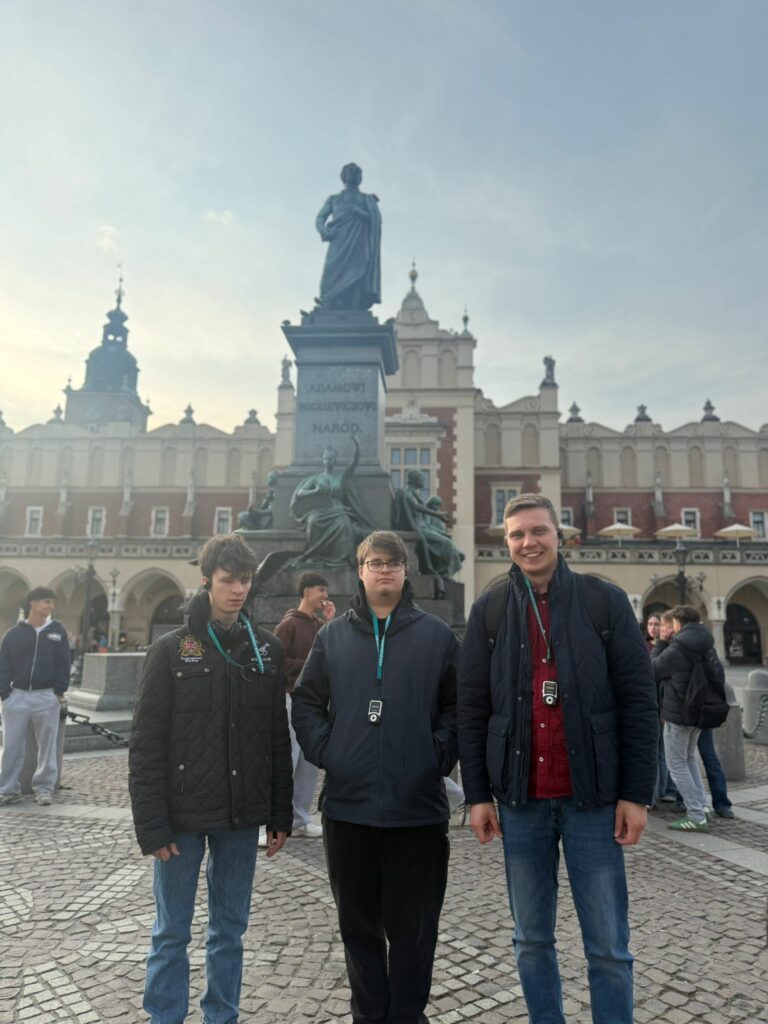 Na  zdjęciu trzej uczestnicy wyjazdu – dwóch maturzystów oraz towarzyszący im opiekun – pozują na krakowskim Rynku Głównym przed pomnikiem Adama Mickiewicza, co było częścią ich programu zwiedzania najważniejszych zabytków miasta.