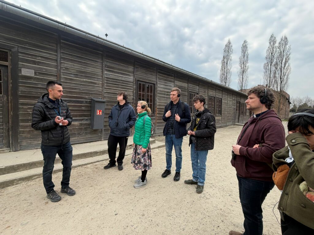 Uczestnicy pielgrzymki w zadumie wsłuchują się w słowa przewodnika przed jednym z drewnianych baraków na terenie Muzeum Auschwitz-Birkenau, co stanowiło kluczowy element ich edukacyjnej podróży poświęconej pamięci i godności człowieka.