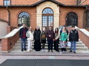 Na zdjęciu grupa maturzystów wraz z opiekunami, w tym księdzem i siostrą zakonną, stoi uśmiechnięta na schodach przed wejściem do Sanktuarium Bożego Miłosierdzia w Krakowie-Łagiewnikach, co stanowiło duchowy punkt ich pielgrzymki połączonej z lekcją historii.