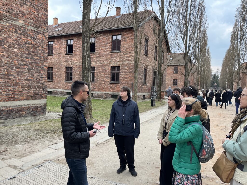 Maturzyści w skupieniu słuchają przewodnika na terenie Miejsca Pamięci i Muzeum Auschwitz-Birkenau. To ważne i trudne doświadczenie, które pozwoliło im na chwilę refleksji nad historią i tragizmem przeszłości. Przejście między historycznymi blokami w milczeniu było jedną z najbardziej poruszających części wyjazdu.