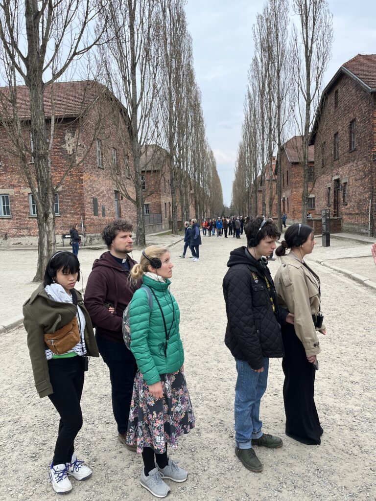 Uczestnicy wyjazdu w milczeniu i pełnym skupieniu przemierzają historyczne alejki obozu Auschwitz I. Słuchawki na uszach pozwalają im w pełni oddać się narracji przewodnika, co w otoczeniu surowych, ceglanych bloków tworzy atmosferę głębokiej zadumy nad losem ofiar. Był to czas osobistej refleksji, który na długo zapadnie w pamięć każdemu z maturzystów.