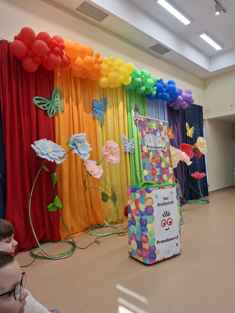 Scena udekorowana kolorowymi balonami, materiałami i kwiatami. Na pierwszym planie napis Pan profesorek