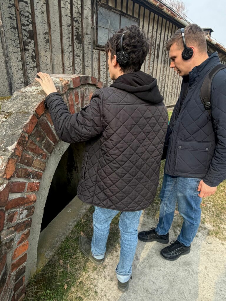 Dwaj uczestnicy wyjazdu, maturzysta i opiekun, w skupieniu oglądają pozostałości obozowej infrastruktury w Muzeum Auschwitz-Birkenau, co stanowiło dla nich przejmującą lekcję o tragicznych wydarzeniach II wojny światowej i cierpieniu drugiego człowieka.