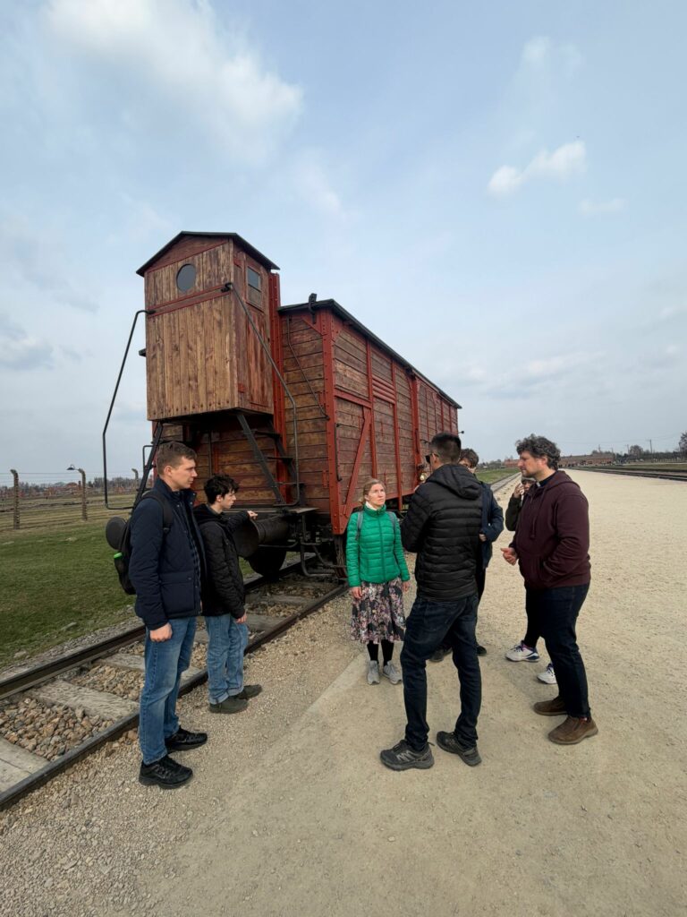 Maturzyści wraz z opiekunami zatrzymali się w zadumie przy historycznym wagonie na rampie w Muzeum Auschwitz-Birkenau, co było poruszającym elementem ich lekcji o tragicznych losach więźniów i poszanowaniu godności ludzkiej.
