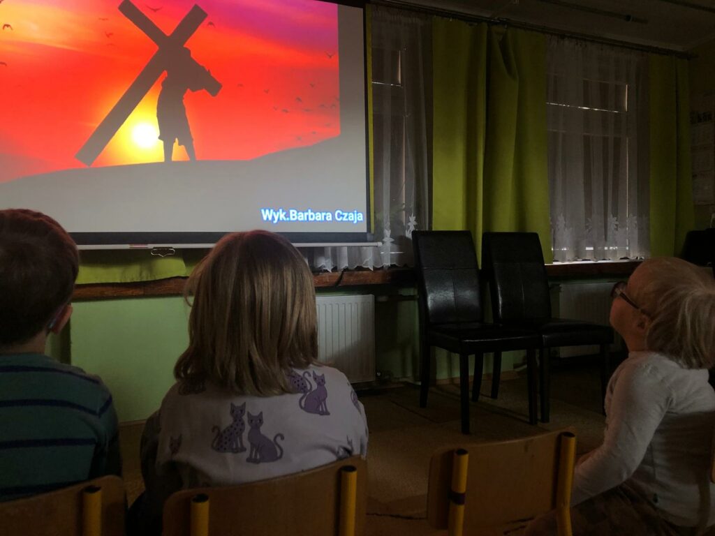 Siedzące dzieci wpatrują się w duży ekran, na nim człowiek niosący krzyż,  na tle zachodzącego słońca