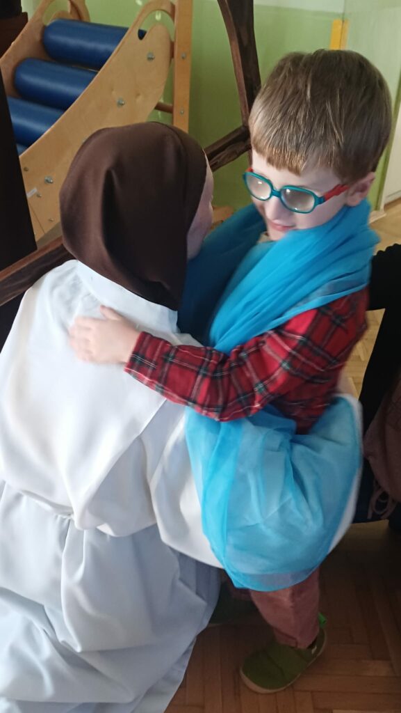 Chłopiec owinięty niebieskim szalem, obejmuje siostrę zakonną podczas odgrywania scenki biblijnej