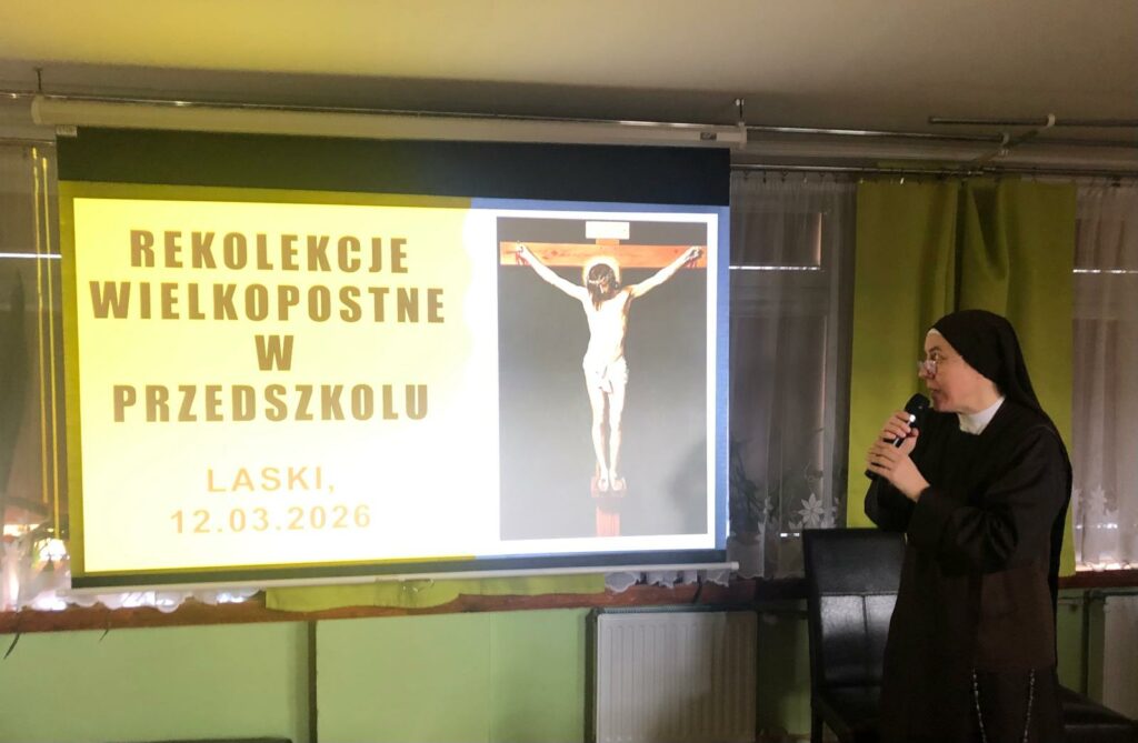 Siostra zakonna trzymająca mikrofon, w tle ekran,  na nim wyświetlony napis:  "Rekolekcje Wielkopostne w przedszkolu" oraz obrazek ukrzyżowanego Pana Jezusa