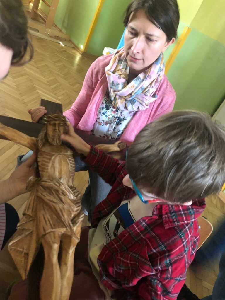 Chłopiec dotyka figury Pana Jezusa na krzyżu, za nim nauczycielka podtrzymuje krzyż