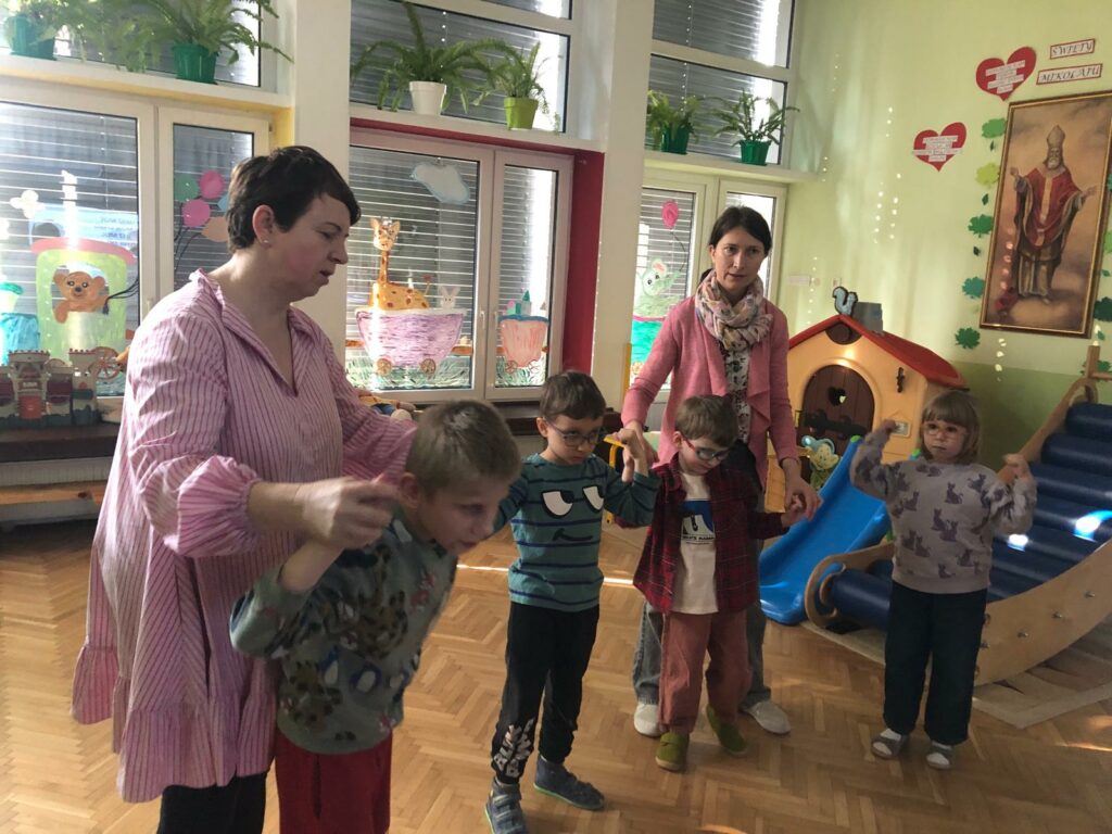 Dzieci stoją z uniesionymi do góry rękami  podczas śpiewu piosenki, za nimi opiekunki