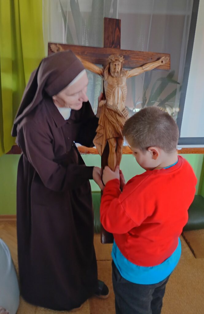 Stojący chłopiec dotyka figury Pana Jezusa na krzyżu, obok siostra zakonna 