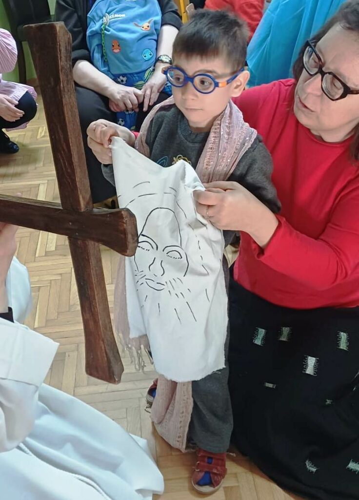 Chłopiec trzyma chustę, na której widać szkic twarzy Pana Jezusa, za nim opiekunka