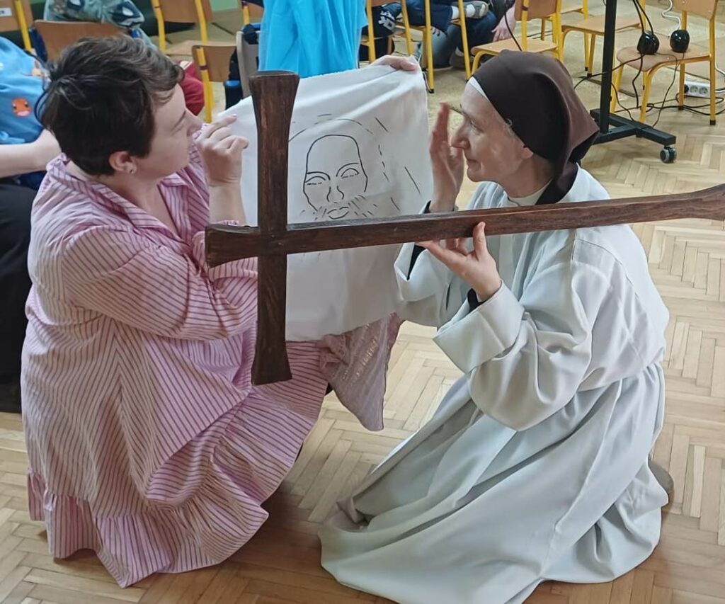 Kobieta trzyma chustę ze  ze szkicem twarzy Pana Jezusa, obok siostra zakonna w albie trzymająca krzyż