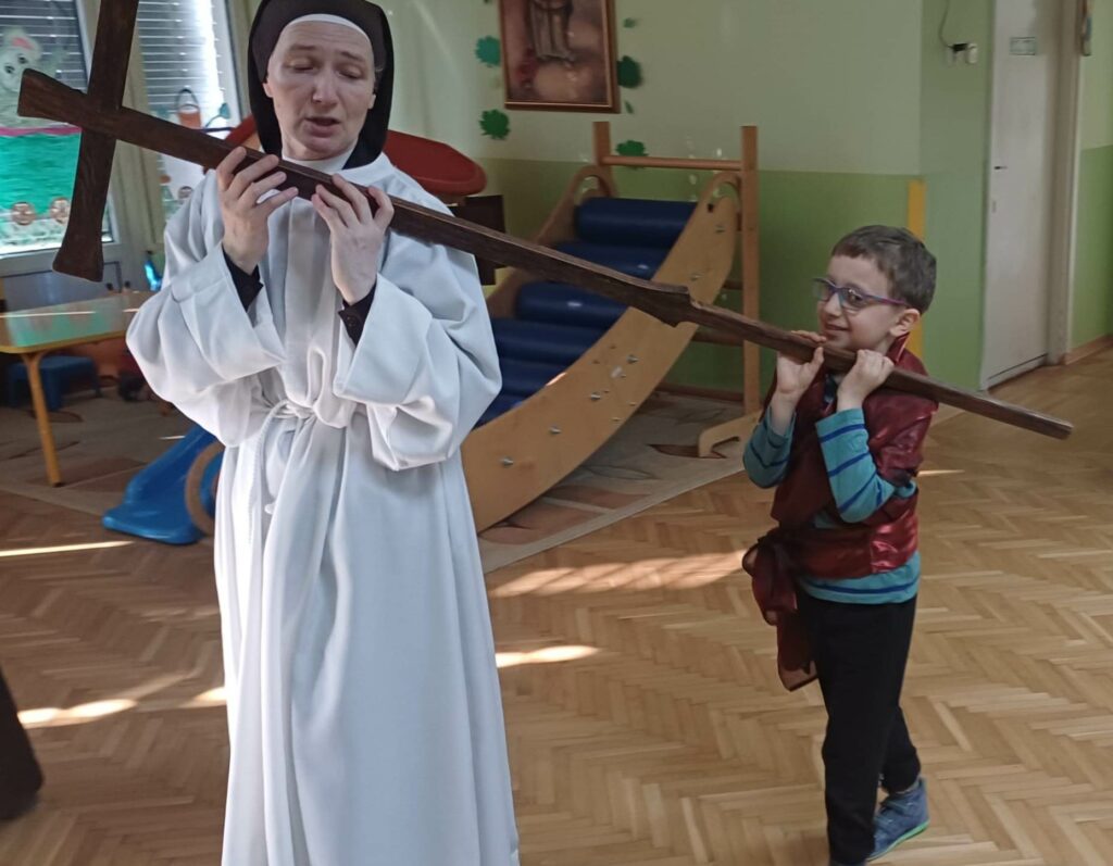 Siostra zakonna w albie, za nią  chłopiec, wspólnie  niosą krzyż, 