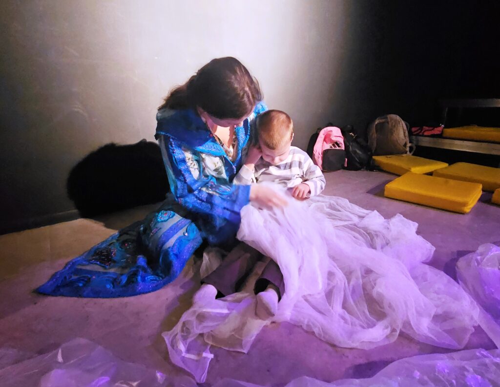 Chłopiec siedzi na podłodze, nogi ma przykryte cienkim materiałem- elementem dekoracji, obok aktorka  